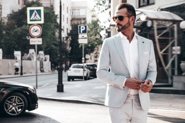 Yakışıklı, kendine güvenen, stil sahibi, lamberseksüel bir modelin portresi. Zarif beyaz takım elbise giymiş modern bir adam. Moda erkeği gün batımında Avrupa şehrinin arka planında poz veriyor. Güneş gözlükleriyle