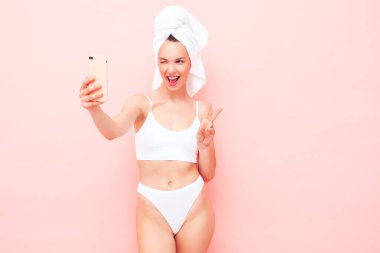 Beyaz iç çamaşırlı, gülümseyen genç bir kadın. İç çamaşırları ve kafasında havluyla stüdyoda pembe duvar pozu veren seksi tasasız manken. Pozitif ve mutlu bir kadın sabah keyfine bakıyor. Selfie çekiyor.