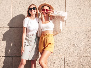 Son moda yaz kıyafetleri içinde iki güzel, gülümseyen hippi kadın. Sokakta beyaz duvarın yanında poz veren kaygısız kadınlar. Pozitif saf modeller gün batımında eğleniyorlar, sarılıyorlar ve deliriyorlar.