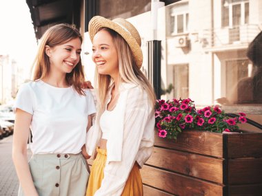 Son moda yaz kıyafetleri içinde iki güzel gülümseyen hippi kadın. Sokakta poz veren kaygısız kadınlar. Pozitif saf modeller gün batımında şapkayla eğleniyorlar, sarılıyorlar ve deliriyorlar.