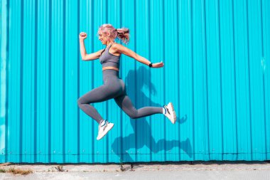 Gri spor elbiseli, pembe saçlı, gülümseyen bir kadın. Mükemmel vücutlu genç, güzel bir manken. Mavi duvarın yanında poz veren bir kadın. Neşeli ve mutlu. Zıpla ve koş.