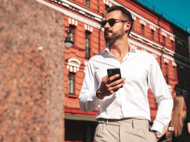 Yakışıklı, kendine güvenen, havalı, havalı, lamberseksüel model. Beyaz gömlek ve pantolon giymiş modern bir adam. Moda erkeği gün batımında Avrupa şehrinde sokakta poz veriyor. Güneş gözlüklü. Akıllı telefona bakıyorum.