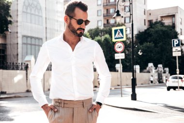 Yakışıklı, kendine güvenen, stil sahibi bir hipster Lambersexual modelin portresi. Beyaz gömlek ve pantolon giymiş modern bir adam. Moda erkeği gün batımında Avrupa şehrinin arka planında poz veriyor. Güneş gözlükleriyle