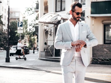 Yakışıklı, kendine güvenen, stil sahibi, lamberseksüel bir modelin portresi. Zarif beyaz takım elbise giymiş modern bir adam. Moda erkeği gün batımında Avrupa şehrinin arka planında poz veriyor. Güneş gözlükleriyle