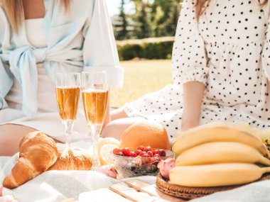 Yaz elbisesi ve şapkası içinde iki güzel, gülümseyen yeniyetme kadın. Dışarıda piknik yapan kaygısız kadınlar. Çimlerin üzerinde oturan, şampanya içen, meyve ve peynir yiyen pozitif modeller.