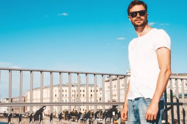 Yakışıklı, gülümseyen hipster Lambersex modelinin portresi. Beyaz tişört giymiş şık bir adam. Moda erkeği mavi gökyüzünün arkasında güneş gözlükleriyle sokak arkasında poz veriyor.