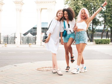 Son moda yaz kıyafetleri içinde üç güzel gülümseyen hippi kadın. Sokakta poz veren tasasız, çok ırklı kadınlar. Güneş gözlükleriyle eğlenen pozitif modeller. Neşeli ve mutlu