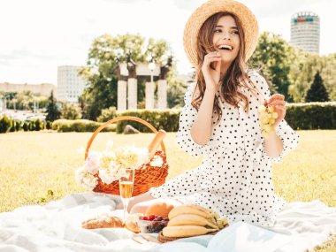 Moda elbisesi ve şapkası olan genç ve güzel hippi kadın. Kaygısız kadın dışarıda piknik yapıyor. Pozitif model çimlerin üzerinde oturuyor, meyve ve peynir yiyor, şampanya içiyor.