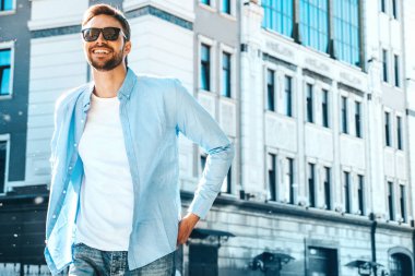 Yakışıklı, güler yüzlü, stil sahibi, lamberseksüel bir modelin portresi. Mavi gömlekli modern bir adam. Moda erkeği, arka planda gökdelenin yanında poz veriyor. Gün batımında dışarıda. 