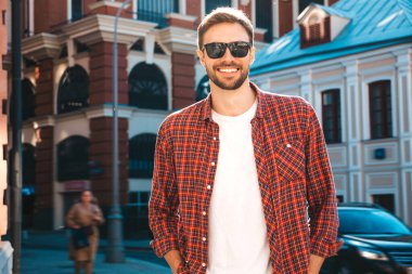Yakışıklı, şık, havalı, havalı, lamberseksüel model. Kırmızı kareli gömlek giymiş modern bir adam. Moda erkeği güneş gözlükleriyle gökdelenin yanında poz veriyor. Gün batımında dışarıda. 