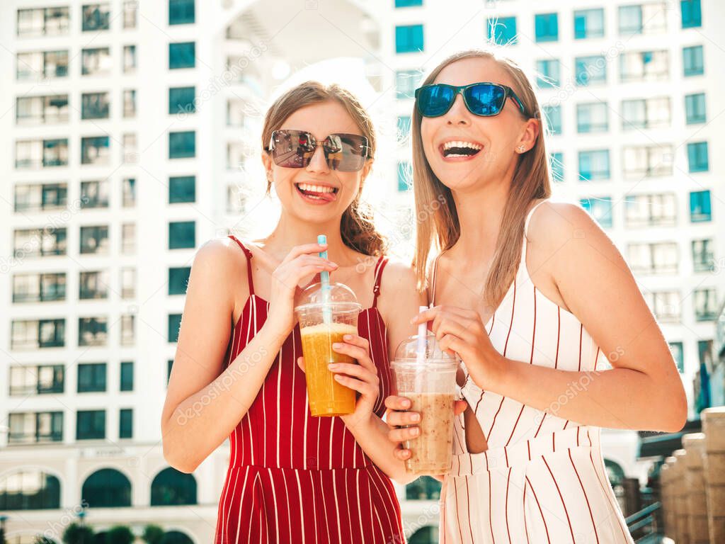 Two young beautiful smiling hipster female in trendy summer clothes. Carefree women posing outdoors.Positive models holding and drinking fresh cocktail smoothie drink in plastic cup with straw