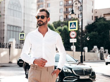 Yakışıklı, kendine güvenen, stil sahibi bir hipster Lambersexual modelin portresi. Beyaz gömlek ve pantolon giymiş modern bir adam. Moda erkeği gün batımında Avrupa şehrinin arka planında poz veriyor. Güneş gözlükleriyle
