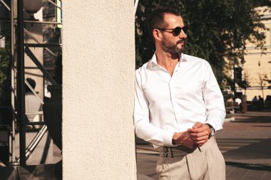 Yakışıklı, kendine güvenen, stil sahibi bir hipster Lambersexual modelin portresi. Beyaz gömlek ve pantolon giymiş modern bir adam. Moda erkeği gün batımında Avrupa şehrinin arka planında poz veriyor. Güneş gözlükleriyle