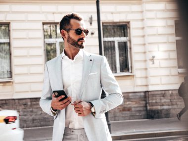 Yakışıklı, kendine güvenen, stil sahibi, lamberseksüel bir modelin portresi. Zarif beyaz takım elbise giymiş modern bir adam. Moda erkeği gün batımında Avrupa şehrinin arka planında poz veriyor. Güneş gözlükleriyle