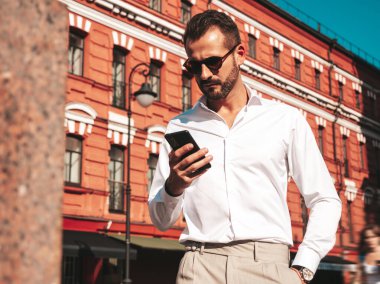 Yakışıklı, kendine güvenen, havalı, havalı, lamberseksüel model. Beyaz gömlek ve pantolon giymiş modern bir adam. Moda erkeği gün batımında Avrupa şehrinde sokakta poz veriyor. Güneş gözlüklü. Akıllı telefona bakıyorum.