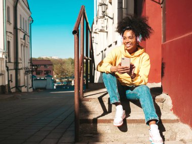 Saçları kıvırcık, güzel siyah bir kadın. Sarı kapüşonlu bir model. Kablosuz kulaklıkla müzik dinlemekten hoşlanan kaygısız bir kadın.