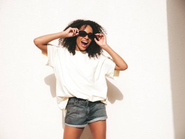 Saçları kıvırcık, güzel siyah bir kadın. Beyaz tişörtlü, hippi bir model. Sokakta güneş gözlüklü beyaz duvarın yanında poz veren kaygısız seksi kadın. Neşeli ve mutlu