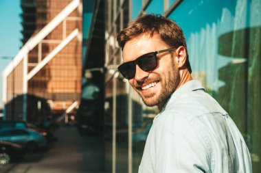 Yakışıklı, güler yüzlü, stil sahibi, lamberseksüel bir modelin portresi. Mavi gömlekli modern bir adam. Moda erkeği, güneş gözlüklü gökdelenlerin yanında sokak arkasında poz veriyor.