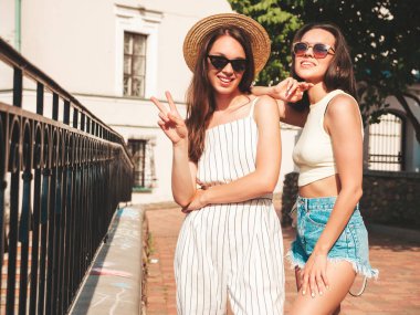 Son moda yaz kıyafetleri içinde iki güzel gülümseyen yeniyetme kadın. Sokakta şapkalı kayla poz veren kaygısız kadınlar. Pozitif saf modeller gün batımında eğleniyorlar, sarılıyorlar. 