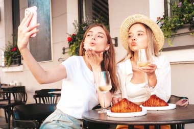 Son moda yaz kıyafetleri içinde gülümseyen iki genç bayan. Sokakta Veranda Kafe 'de poz veren kaygısız kadınlar. Beyaz şarap içen, kruvasan yiyen, selfie çeken pozitifler.