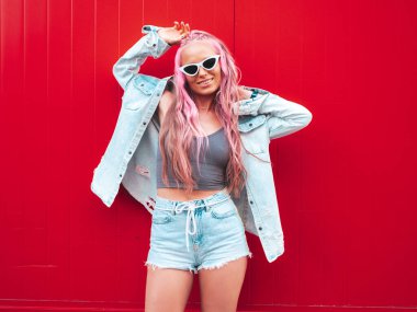 Genç, güzel, gülümseyen bir hippi kadın, son moda yaz kot şortuyla, pembe saç stiliyle. Sokakta kırmızı duvarın yanında poz veren kaygısız seksi bir model. Güneş gözlüklü pozitif model.