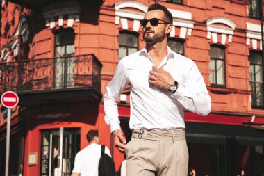 Yakışıklı, kendine güvenen, stil sahibi bir hipster Lambersexual modelin portresi. Beyaz gömlek ve pantolon giymiş modern bir adam. Moda erkeği gün batımında Avrupa şehrinin arka planında poz veriyor. Güneş gözlükleriyle