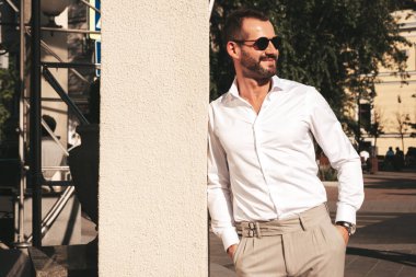 Yakışıklı, kendine güvenen, stil sahibi bir hipster Lambersexual modelin portresi. Beyaz gömlek ve pantolon giymiş modern bir adam. Moda erkeği gün batımında Avrupa şehrinin arka planında poz veriyor. Güneş gözlükleriyle