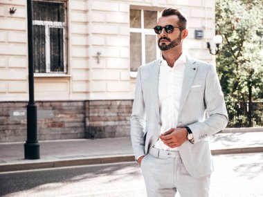 Yakışıklı, kendine güvenen, stil sahibi, lamberseksüel bir modelin portresi. Zarif beyaz takım elbise giymiş modern bir adam. Moda erkeği gün batımında Avrupa şehrinin arka planında poz veriyor. Güneş gözlükleriyle