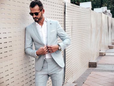 Yakışıklı, kendine güvenen, stil sahibi, lamberseksüel bir modelin portresi. Zarif beyaz takım elbise giymiş modern bir adam. Moda erkeği gün batımında çitlerin yanında poz veriyor. Güneş gözlükleriyle