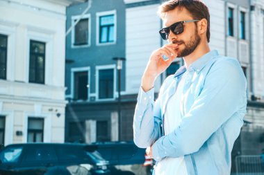 Yakışıklı, kendine güvenen, havalı, havalı, lamberseksüel model. Mavi gömlekli, modern bir adam. Moda erkeği güneş gözlükleriyle gökdelenin yanında poz veriyor. Düşünce. Kollar çapraz.