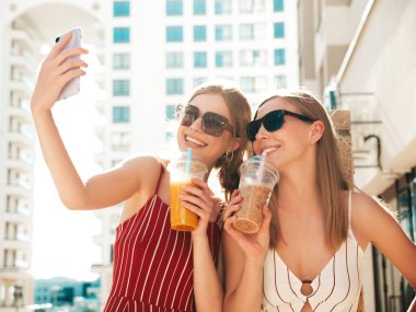 Moda kıyafetleri içinde gülümseyen iki güzel hippi kadın. Dışarıda poz veren kaygısız kadınlar. Pozitif modeller. Pipetle taze kokteyl içecekleri içiyorlar.