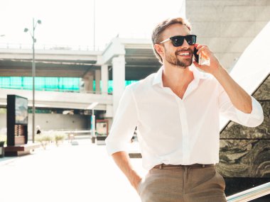 Yakışıklı, güler yüzlü, stil sahibi, lamberseksüel model. Beyaz gömlekli, modern bir adam. Sokakta güneş gözlükleriyle poz veren modacı erkek. Gün batımında dışarıda. Akıllı telefon kullanarak. Konuşarak.