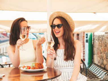 Son moda yaz kıyafetleri içinde iki güzel gülümseyen yeniyetme kadın. Sokakta Veranda Kafe 'de poz veren kaygısız kadınlar. Şapkada beyaz şarap içen pozitif modeller.