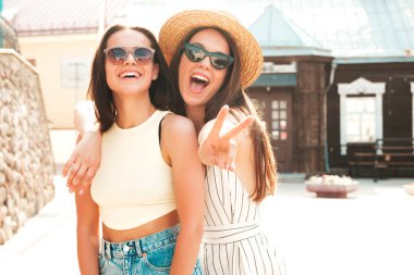 Son moda yaz kıyafetleri içinde iki güzel gülümseyen yeniyetme kadın. Sokakta şapkalı kayla poz veren kaygısız kadınlar. Pozitif saf modeller gün batımında eğleniyorlar, sarılıyorlar. 