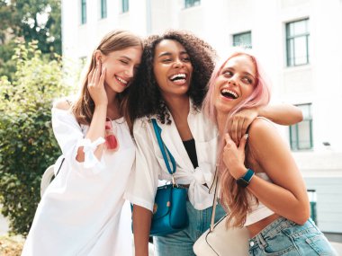 Son moda yaz kıyafetleri içinde üç güzel gülümseyen hippi kadın. Sokakta poz veren tasasız, çok ırklı kadınlar. Güneş gözlükleriyle eğlenen pozitif modeller. Neşeli ve mutlu