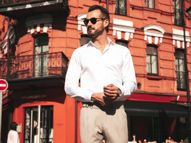 Yakışıklı, kendine güvenen, stil sahibi bir hipster Lambersexual modelin portresi. Beyaz gömlek ve pantolon giymiş modern bir adam. Moda erkeği gün batımında Avrupa şehrinin arka planında poz veriyor. Güneş gözlükleriyle