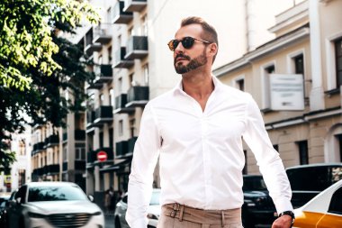 Yakışıklı, kendine güvenen, stil sahibi bir hipster Lambersexual modelin portresi. Beyaz gömlek ve pantolon giymiş modern bir adam. Moda erkeği gün batımında Avrupa şehrinin arka planında poz veriyor. Güneş gözlükleriyle