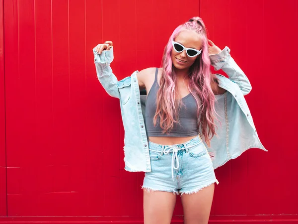 ピンクの髪型で流行の夏のジーンズのショートパンツで若い美しい笑顔ヒップスター女性 赤い壁の近くの通りにポーズを取るセクシーな屈託のないモデル サングラスで肯定的なモデル屋外 — ストック写真