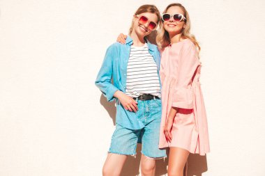Son moda yaz kıyafetleri ve elbiseleri içinde iki güzel, gülümseyen hippi kadın. Sokakta beyaz duvarın yanında poz veren kaygısız kadınlar. Pozitif saf modeller eğleniyor, sarılıyor ve deliriyor.