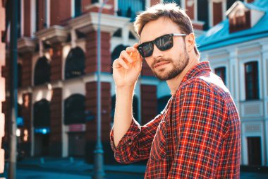 Yakışıklı, şık, havalı, havalı, lamberseksüel model. Kırmızı kareli gömlek giymiş modern bir adam. Moda erkeği güneş gözlükleriyle gökdelenin yanında poz veriyor. Gün batımında dışarıda. 