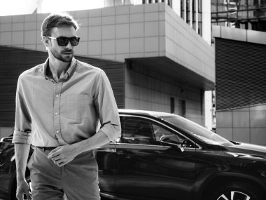 Yakışıklı, kendine güvenen, stil sahibi bir hipster Lambersexual modelin portresi. Gömlek giymiş modern bir adam. Moda erkek, güneş gözlüklü gökdelenlerin yanında sokak arkasında poz veriyor. Lüks bir arabanın yanında.