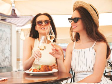 Son moda yaz kıyafetleri içinde iki güzel gülümseyen yeniyetme kadın. Sokakta Veranda Kafe 'de poz veren kaygısız kadınlar. Şapkada beyaz şarap içen pozitif modeller.