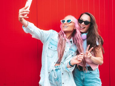 Son moda yaz kıyafetleri içinde iki güzel gülümseyen yeniyetme kadın. Sokakta pembe saçlı, kırmızı duvarın yanında poz veren kaygısız kadınlar. Pozitif saf modeller eğleniyor. Neşeli ve mutlu. Selfie çekiyor.