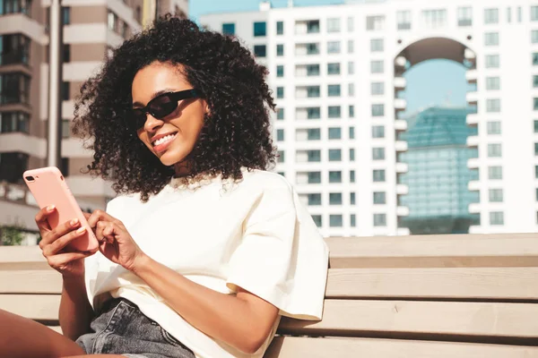 Beautiful black woman with afro curls hairstyle.Smiling model in white hoodie. Sexy carefree female posing on the street background in sunglasses. Looking at smartphone screen, using apps