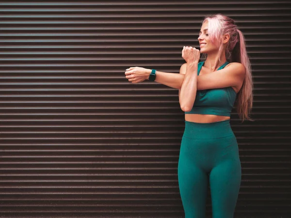 Fitness smiling woman in green sports clothing with pink hair. Young beautiful model with perfect body.Female posing in the street near roller shutter wall. Stretching out before training