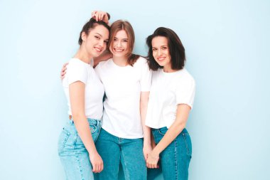 Aynı yaz moda olan beyaz tişört ve kot pantolon giyen üç güzel, gülümseyen hippi kadın. Stüdyoda açık mavi duvarın yanında poz veren kaygısız seksi kadınlar. Neşeli ve pozitif modeller eğleniyor.