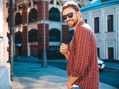 Yakışıklı, şık, havalı, havalı, lamberseksüel model. Kırmızı kareli gömlek giymiş modern bir adam. Moda erkeği güneş gözlükleriyle gökdelenin yanında poz veriyor. Gün batımında dışarıda. 