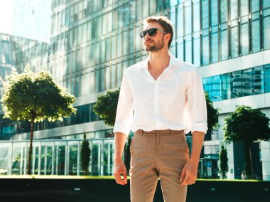 Yakışıklı, kendine güvenen, stil sahibi, Lambersex mankeni portresi. Beyaz gömlek giymiş modern bir adam. Moda erkeği, güneş gözlüklü gökdelenlerin yanında sokak arkasında poz veriyor.