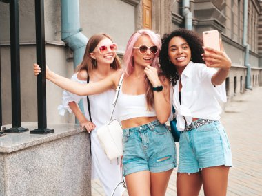 Son moda yaz kıyafetleri içinde üç güzel gülümseyen hippi kadın. Sokakta poz veren tasasız, çok ırklı kadınlar. Güneş gözlükleriyle eğlenen pozitif modeller. Selfie çekiyoruz.