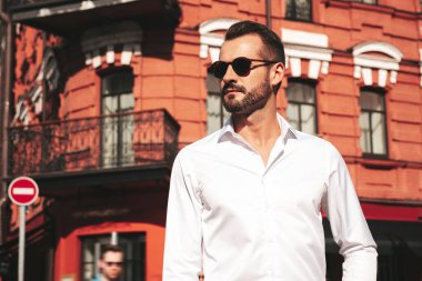 Yakışıklı, kendine güvenen, stil sahibi bir hipster Lambersexual modelin portresi. Beyaz gömlek ve pantolon giymiş modern bir adam. Moda erkeği gün batımında Avrupa şehrinin arka planında poz veriyor. Güneş gözlükleriyle